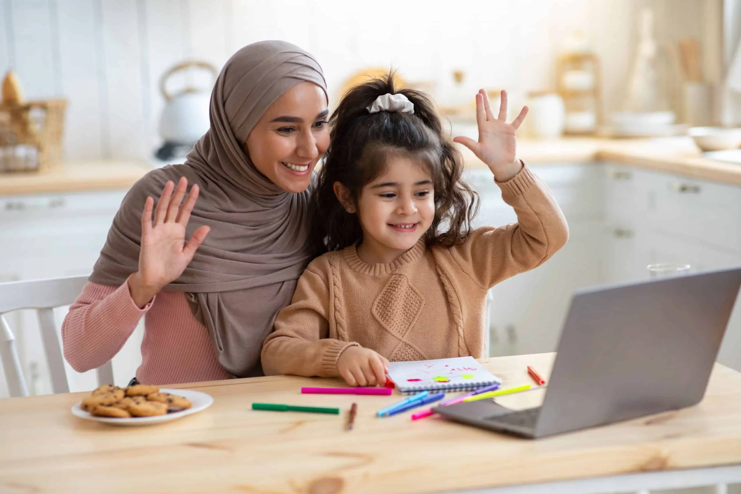 Mother in hijab and young daughter waving during a video call at home