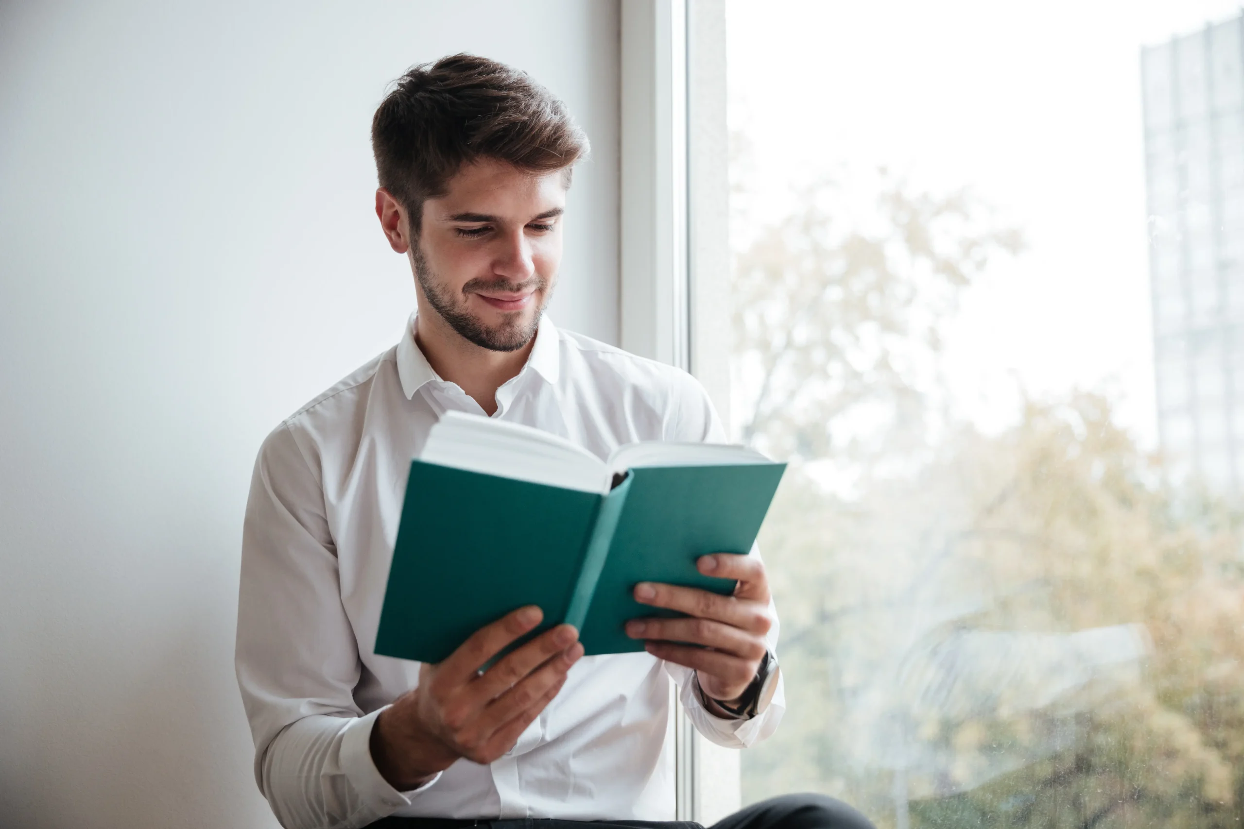 Man in a white shirt reading a book by a window