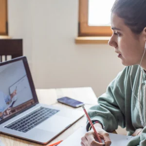 Woman taking notes while attending an online Arabic lesson with a tutor visible on her laptop screen