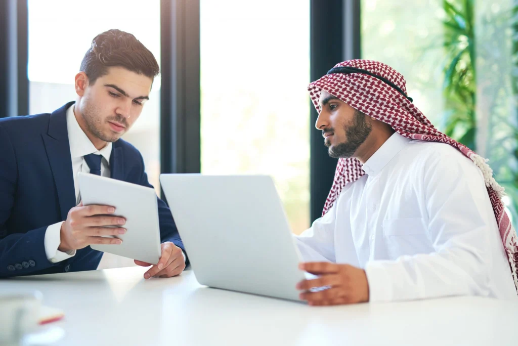 Two businessmen, one in a suit and one in traditional Gulf dress, reviewing data together on a laptop and tablet