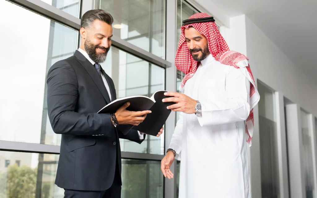 Businessman in a suit reviewing a document with an Arab business partner in traditional dress in an office hallway