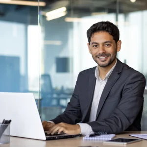 Smiling professional in a business suit working on a laptop in a modern glass-walled office, representing career growth through a business Arabic course