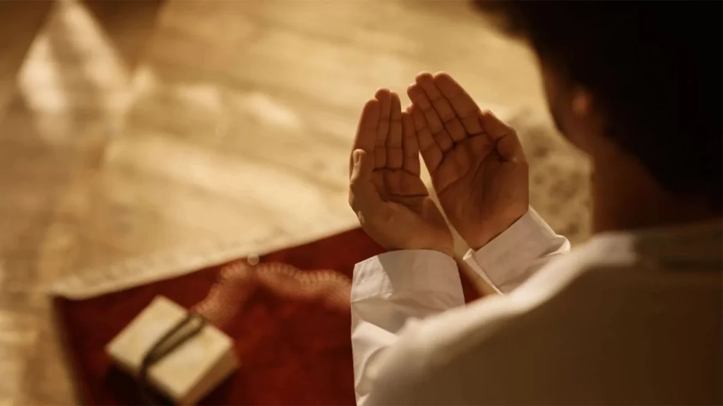 Man raising his hands in dua over a prayer mat with prayer beads and a small book nearby