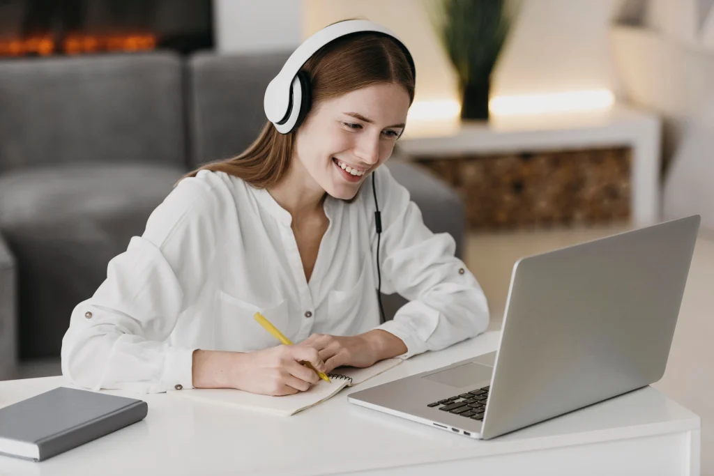 Smiling woman wearing headphones taking notes during an online lesson on her laptop